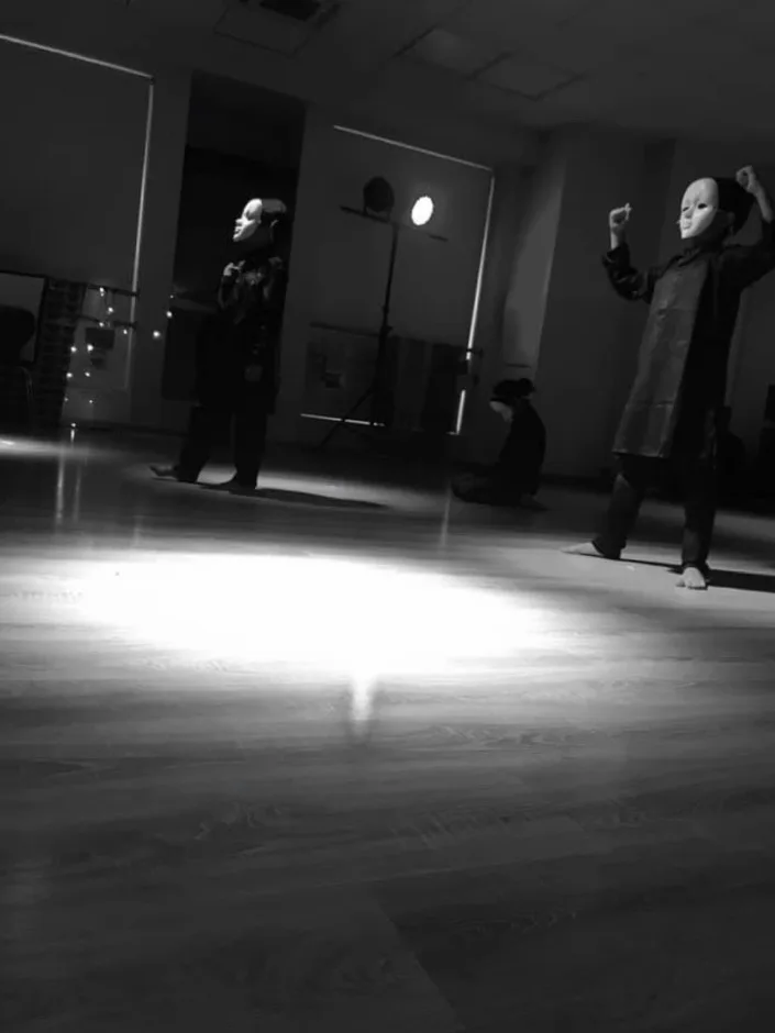Performers in white masks rehearsing in a darkened studio